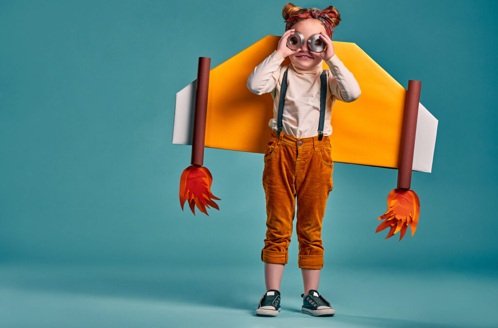 Smiling child with paper plane wings and eyeglasses isolated on blue.